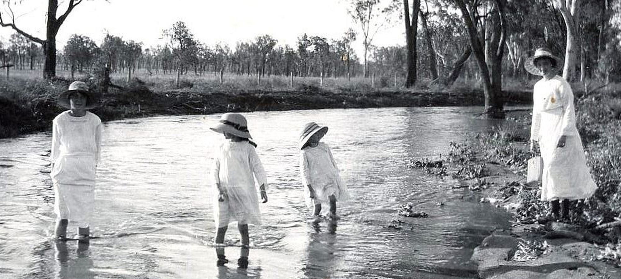 Images from Early Banana, Miller Family. A black and white image of 3 girls wading in water with a woman standing on the banks.